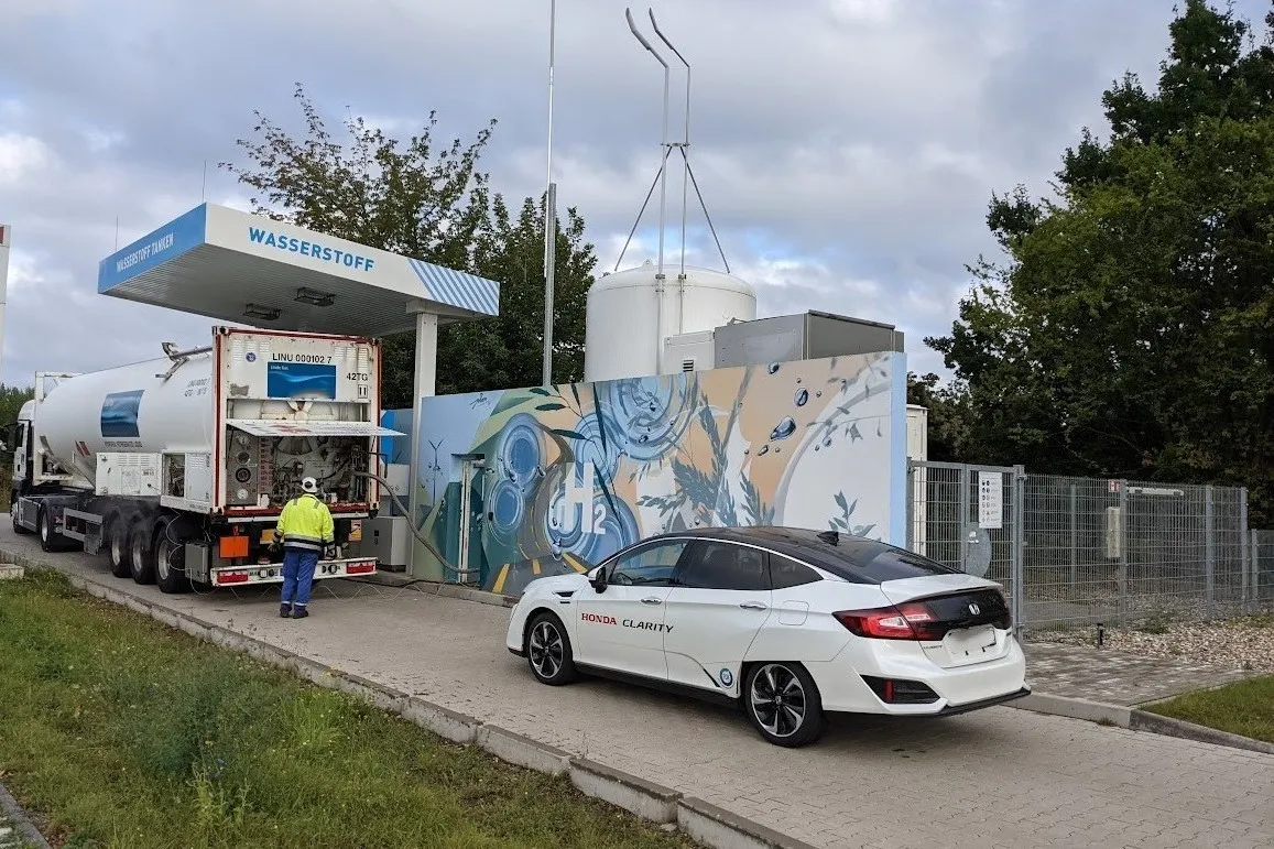 Tanker trailer unloading liquid hydrogen at a refueling station, with a fuel cell car parked at the dispenser